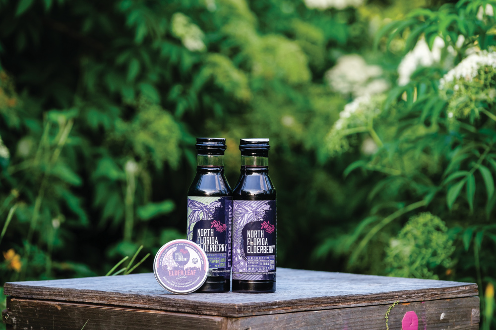 NEF Product image; 2 Elderberry Pure Juice bottles and a leaf salve container. Outside on a wooden table amongst elderberry bushes